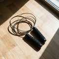 Corde à sauter Amplitude posée sur un parquet : câble acier lové et deux poignées noires